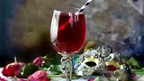 Dried red rose petals and a steampunk mask surround a glass goblet of crimson zombie cocktail that's low carb.