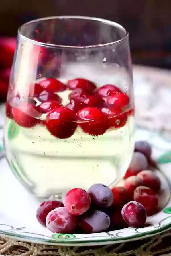 Close up of low carb white wine spritzer in a clear glass with frozen cranberries floating in it and frosted cranberries at the base