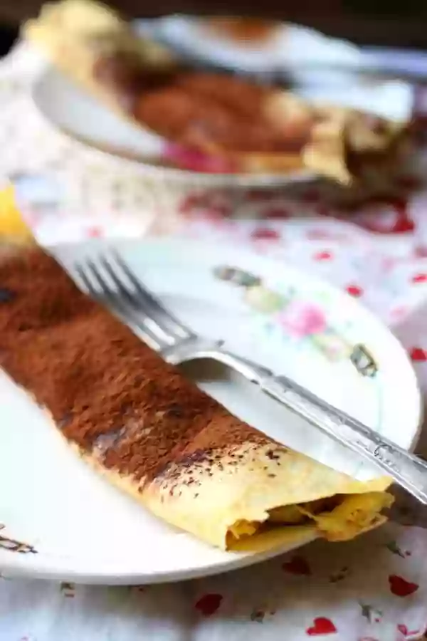 Low carb chocolate mousse crepes are dusted with cocoa and place on a white plate with a rose pattern. Plate sits on a pink patterned tablecloth