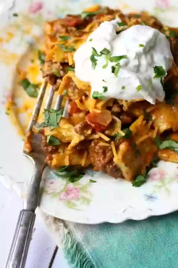 A silver fork is scooping up some of the low carb taco casserole. The tex mex casserole is on a vintage white plate with flowers on it. a greenish colored napkin is under the plate