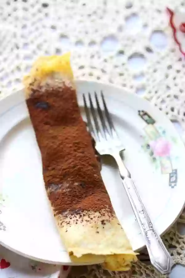 looking down on a low carb chocolate mousse crepe on a white plate with a rose pattern on the edge. A silver fork is on the side. Plate is on a lace tablecloth