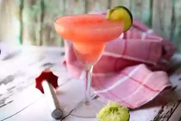 Skinny Watermelon Margarita in a glass on a chipped painted white table. A Red checked towel is tossed on to the table and a squeezed lime half is in the forground