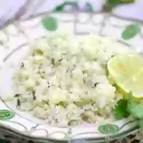 A dish of cilantro lime cauliflower rice is places on a wood table and garnished with a slice of bright green lime.