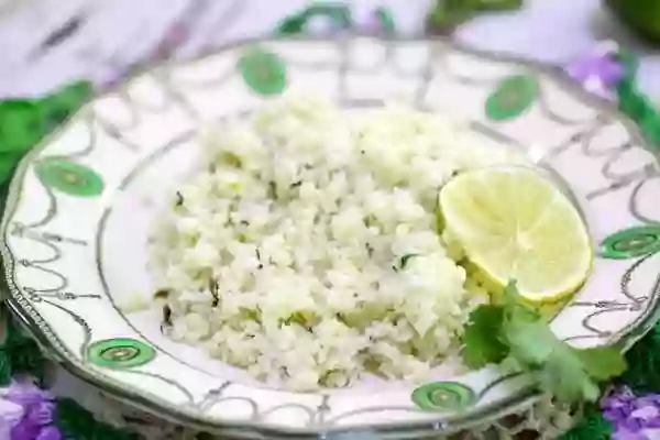 A dish of cilantro lime cauliflower rice is places on a wood table and garnished with a slice of bright green lime.