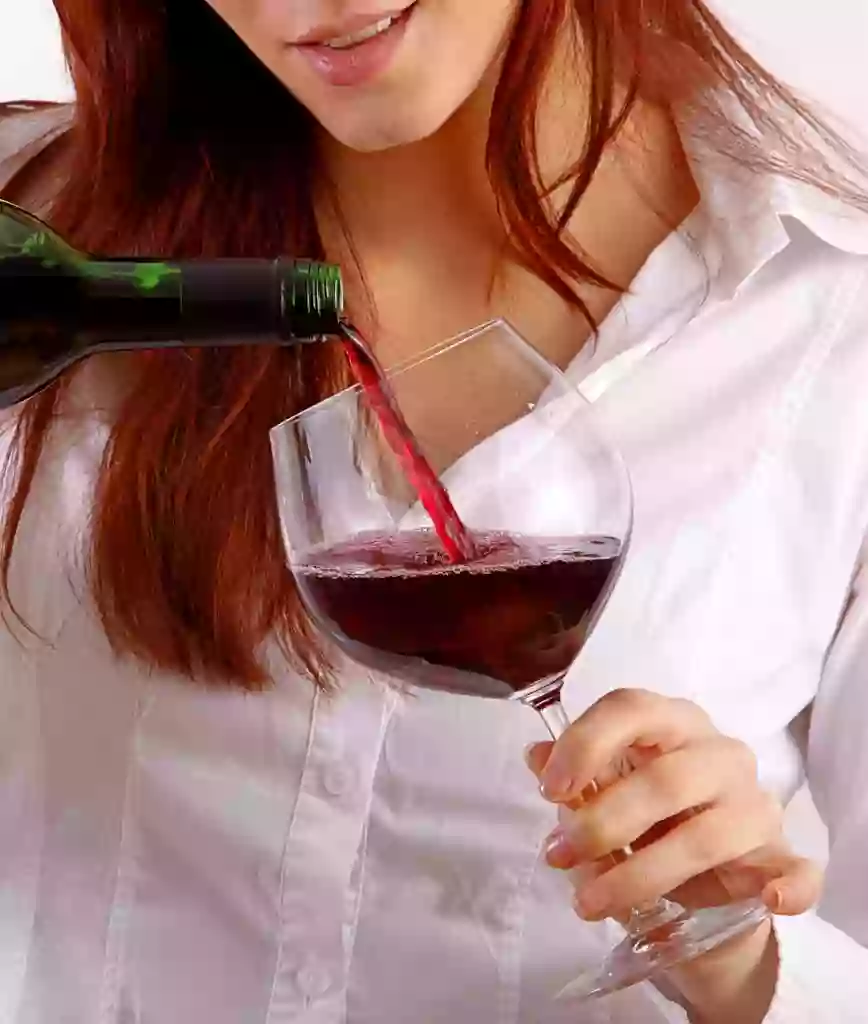 A women in a white shirt with red hair pouring a glass of red wine into a wine glass she is holding in her left hand