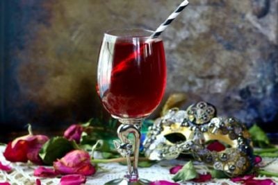 Dried red rose petals and a steampunk mask surround a glass goblet of crimson zombie cocktail that's low carb.