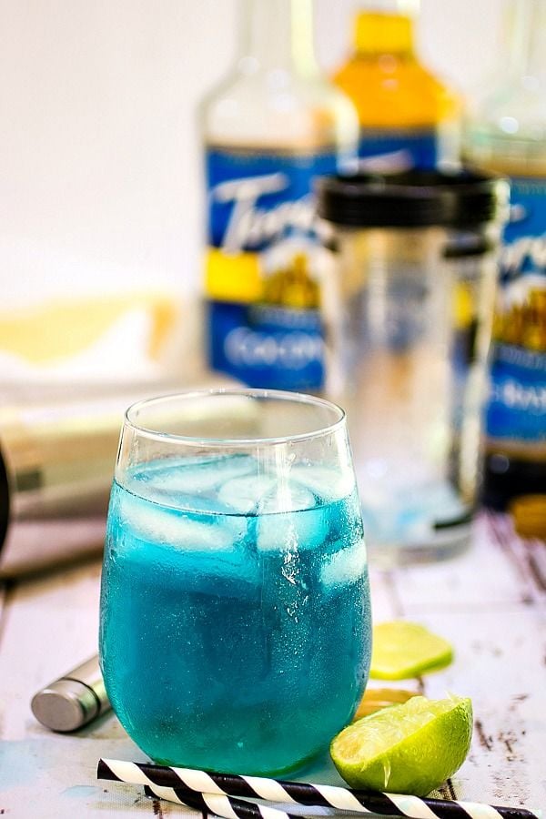 A blue lagoon cocktail with bottles of Torani syrups in the background